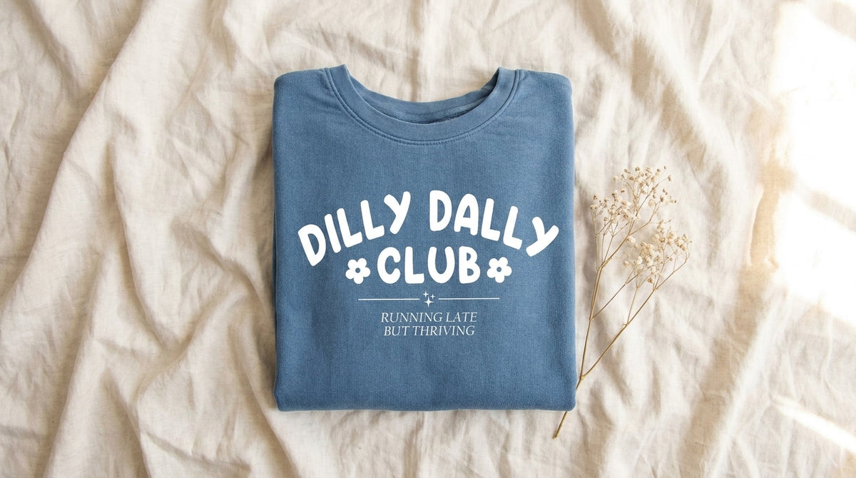 Dilly Dally Club blue sweatshirt folded on rumpled ivory linen with baby's breath – Peachy Sunday gentle productivity guide