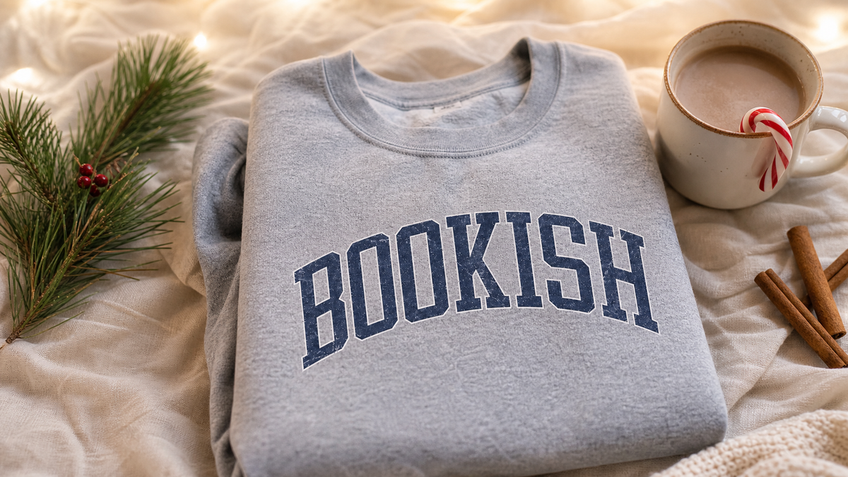 Cozy December holiday flat lay with a Bookish sweatshirt, mug with candy cane, pine sprig, and cinnamon sticks on cream linen