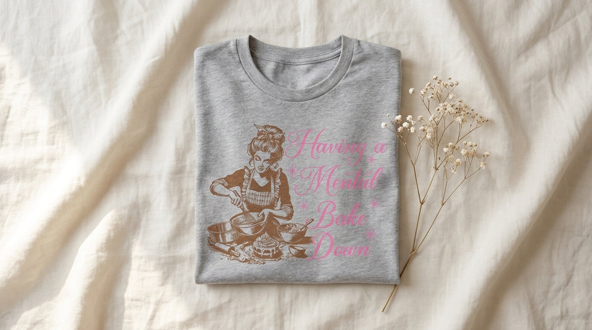 Folded heather grey Having a Mental Bake Down graphic tee on rumpled ivory linen with baby's breath sprig and soft window light – Peachy Sunday holiday cookie prep guide