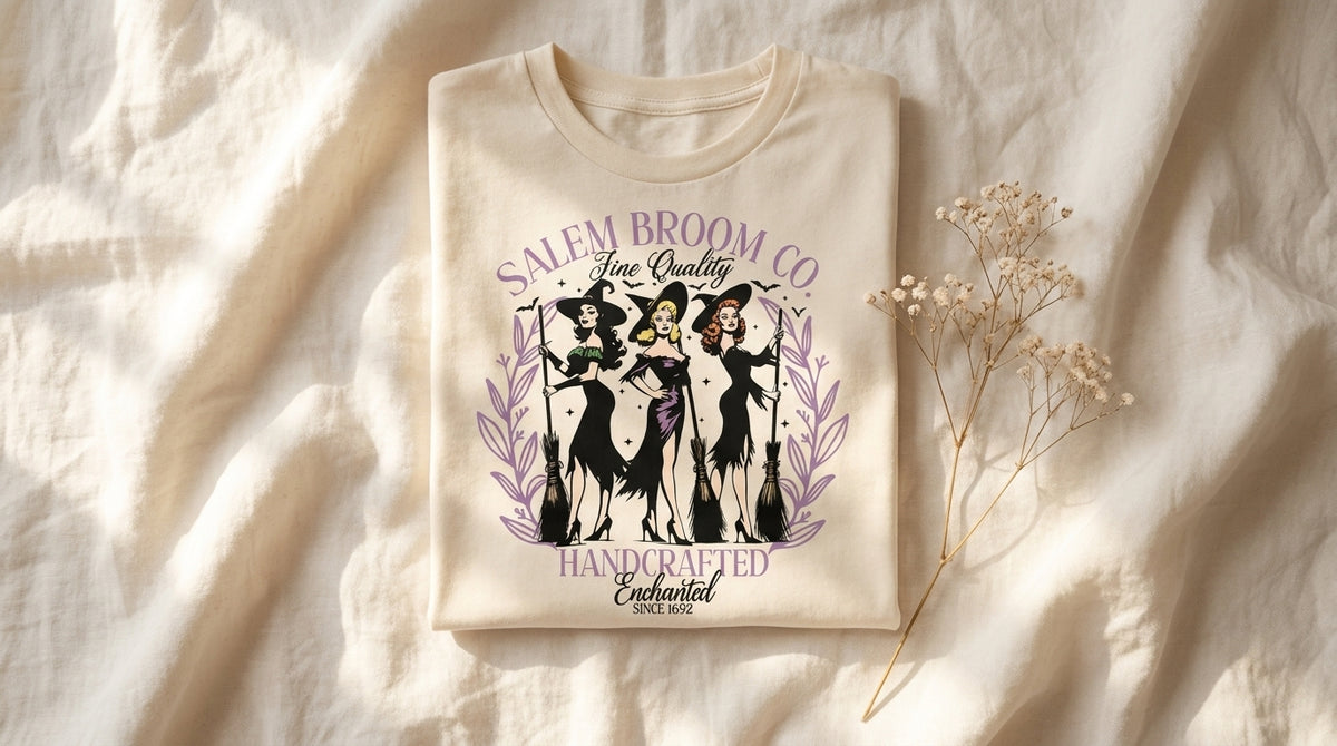 Folded cream Salem Broom Co graphic tee on rumpled ivory linen with baby's breath sprig