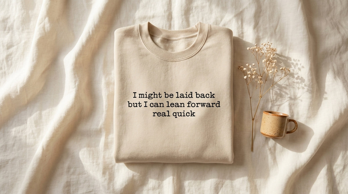 Folded sand I Might Be Laid Back sweatshirt on rumpled ivory linen with window light and baby's breath sprig