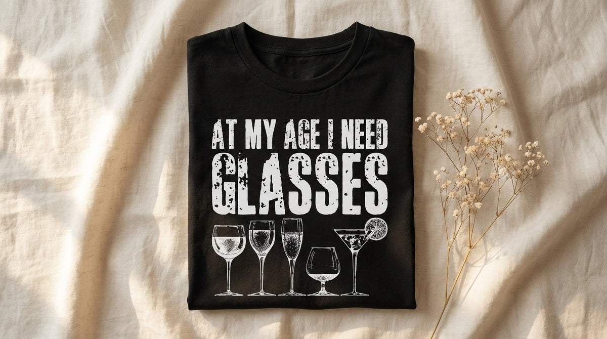 At My Age I Need Glasses graphic tee neatly folded on rumpled ivory linen with baby's breath sprig and morning window light – Peachy Sunday milestone birthday gift guide