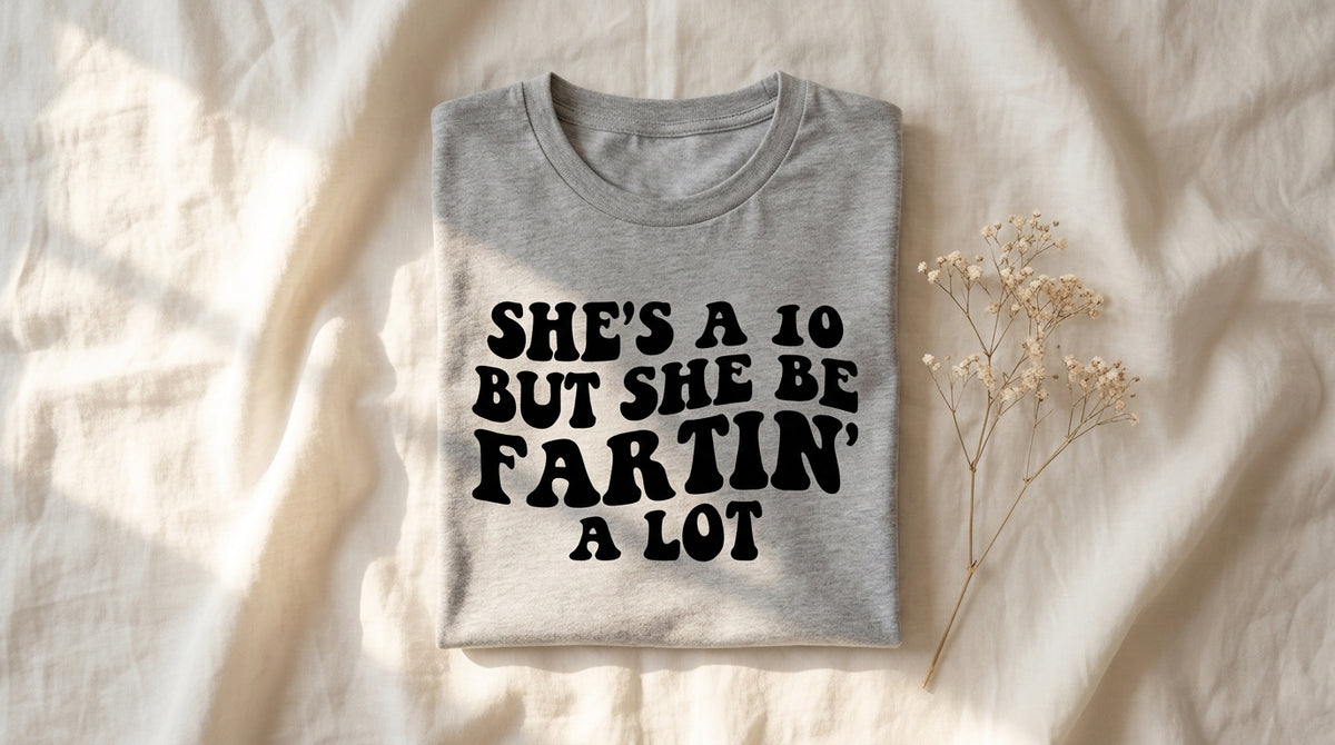 She's A 10 But She Be Fartin A Lot graphic tee folded on rumpled ivory linen with baby's breath – Peachy Sunday meme explainer